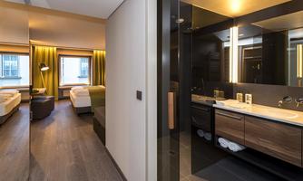 A modern hotel room with two beds and large windows. In the background, there is an elegant bathroom with a sink and shower.