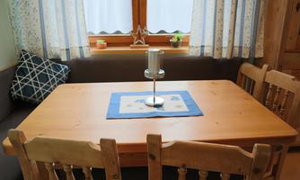 A cozy dining area with a wooden table and chairs. A blue table runner with a candle is on the table, and the windows are adorned with light curtains.