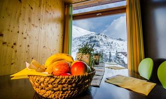 A basket of fresh fruit is on a table in a cozy room. Through the window, a snowy mountain landscape can be seen.