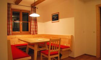 A cozy dining area with a wooden bench and a table. The walls are light, and the window has checkered red curtains.