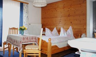 A cozy room with a bed and a dining table. The walls have a wooden look, and the windows let in plenty of light.