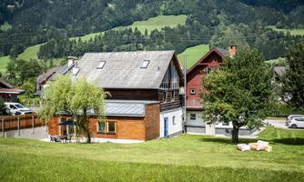 A modern house with wooden cladding and a large garden. In the background, green hills and trees can be seen.
