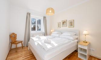 Consistent bedroom with a large bed, white bedding, and a bedside table. The room has light walls, wooden flooring, and a large window front with curtains.