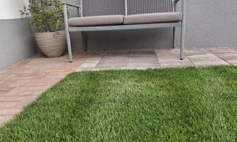 A modern bench stands on a paved floor next to a green lawn. In the background, a gray wall can be seen.