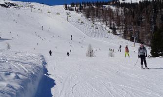 A wide, snow-covered slope with many skiers. In the background, clear blue skies and snow-covered mountains can be seen.
