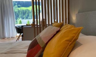 A stylish bedroom with a cozy bed and colorful cushions. In the background, there are large windows overlooking the green landscape.