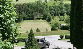 A green landscape with gentle hills and forests. In the foreground, a vehicle is on a quiet road.