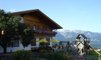 A cozy house with a wooden balcony and colorful flowers. In the foreground, there is a playground and in the background, the mountains stretch out.