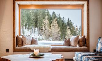 A cozy seating area in front of a large window overlooking a wintry forest. The decor is modern and rustic, with cushions and a lovely table.