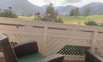 A balcony with two cozy chairs and blooming geraniums. In the background, green meadows and mountains can be seen under a cloudy sky.