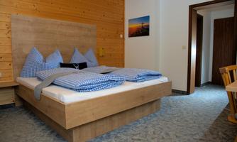 A cozy bedroom with a large bed and checkered bedding. The walls are made of wood, and there is a small table as well as a chair in the room.