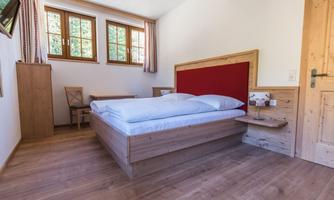 A simple bedroom with a wooden bed and a red headboard. There are windows looking outside and a small table with a chair.