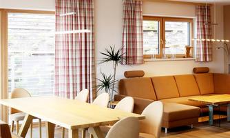 A bright and modern kitchen-living area with a large dining table and a cozy couch. The windows let in plenty of light, and the decoration is simple and stylish.