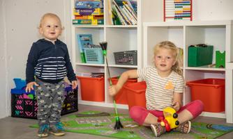 Two children are playing in a room. The boy is standing with a smile, while the girl is busy on the floor with toys.