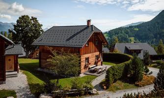 A rustic wooden house surrounded by green landscapes and mountains. The surroundings are calm and inviting, perfect for a relaxing stay.