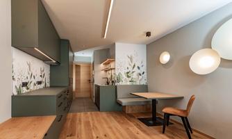 A modern kitchen with green cabinets and wooden flooring. The dining area is equipped with a cozy bench and a table, surrounded by stylish wall designs.