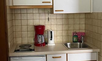 A small kitchenette with white cabinets, a stove, a refrigerator, and a sink. On the countertop, there is a coffee machine and a kettle.
