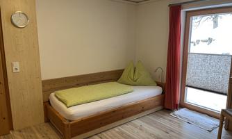 A simple room with a single bed and wooden interior. A balcony with a view is visible through the window.
