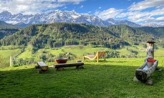 A picturesque landscape with green meadows and snow-capped mountains in the background. In the foreground, there are wooden seating areas.