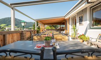 A cozy terrace with tables and chairs, surrounded by plants. In the background, green hills are visible under a clear blue sky.