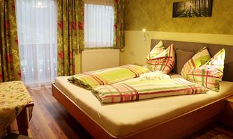A cozy bedroom with a large bed and colorful pillows. The window curtains are floral and the room has a warm, inviting atmosphere.