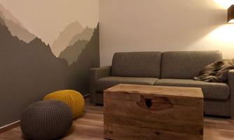 A cozy living room with a gray sofa and a wooden crate. The wall is creatively designed with mountain motifs, and there are two colorful poufs in yellow and gray.