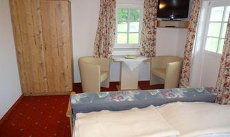 A cozy room with a double bed, two chairs, and a small table. Large windows provide plenty of light, and a TV is also available.