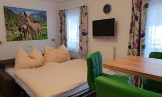 A cozy room with a bed, a table, and brightly green chairs. On the wall hangs a picture of donkeys in a landscape.