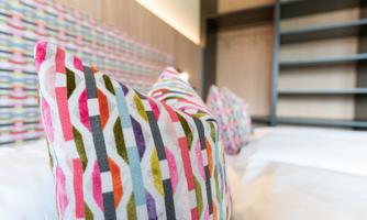 Colorful cushions on a bed in a modern room. The background features a simple wall with a graphic pattern.