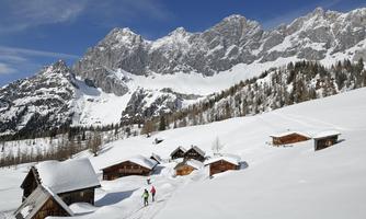 A snowy mountain landscape with traditional wooden houses. In the background, majestic mountains rise under a clear blue sky.