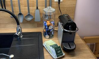 A modern kitchen area with a Nespresso machine and coffee capsules in a glass container. Kitchen utensils are hanging on the wall above the black material sink.