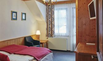 A cozy bedroom with a double bed and an armchair. The walls are painted in light tones, and there is a small table as well as a television.
