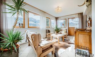 A bright living room with large windows and a cozy decor. Plants bring life to the space and the floor is tiled.