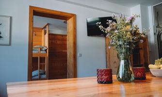 A cozy room with a wooden table and a flower vase. In the background, a television and a bedroom can be seen.