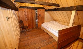 A cozy room with wooden paneling and a single bed. A wooden bench and a door lead to another room.