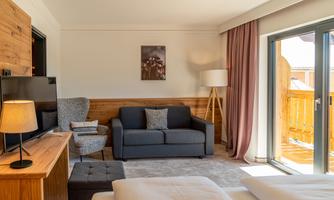 A bright and modern hotel room with a gray sofa, a television, and a table. The room has large windows overlooking a balcony.