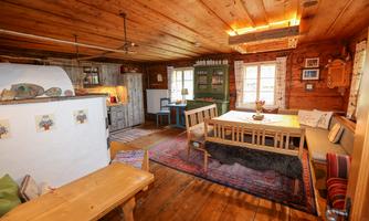A cozy room with wooden ceilings and rustic furnishings. There is a dining area, a kitchen, and large windows that provide plenty of light.