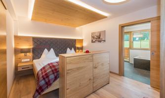 A modern bedroom with wooden details and a cozy bed. Large windows offer a view of the outdoors and plenty of daylight.