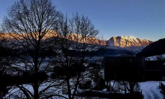 A picturesque winter landscape with snow-covered mountains and bare trees. The sun illuminates the peaks in warm colors.