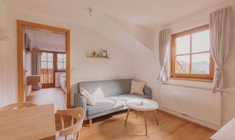A bright and cozy living room with a gray sofa and a small table. Lots of light comes in through the window, and you have a view into an adjacent room.