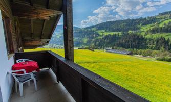 A balcony with a table and a chair that overlooks a vast green meadow with mountains in the background. The sky is blue with some clouds.