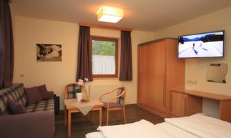 A cozy room with a double bed, a table, and two chairs. A television hangs on the wall and a window lets in daylight.