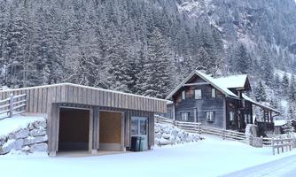 A snowy winter landscape with a wooden house and a shelter. Surrounded by pine trees and mountains.