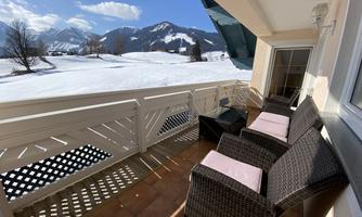 A cozy terrace with rattan furniture that offers a view of snow-covered mountains. The sky is clear and blue.
