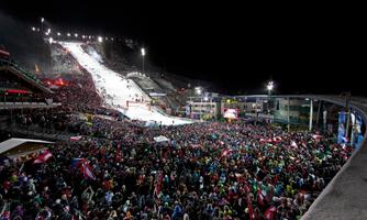 A large crowd gathers at a nighttime sporting event on a lit ski slope. Many spectators hold flags and cheer for the athletes.