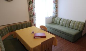 A cozy living area with a yellow tablecloth and a wooden table. There is a seating area with striped cushions and plenty of natural light coming through the window.