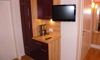 A modern kitchenette with wooden and dark red cabinets. A television hangs on the wall above a countertop with a coffee machine.