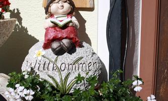 A decorative garden figure sits on a stone with the inscription "Welcome". Surrounded by beautiful flowers and a rustic background.