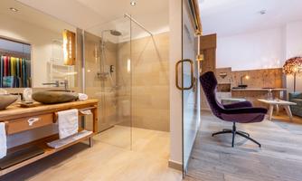 A modern bathroom with a glass shower and a wooden washbasin. In the background, a cozy seating area can be seen.
