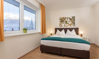 A bright bedroom with a large window overlooking a church. The bed is cozy with white pillows and a green blanket.